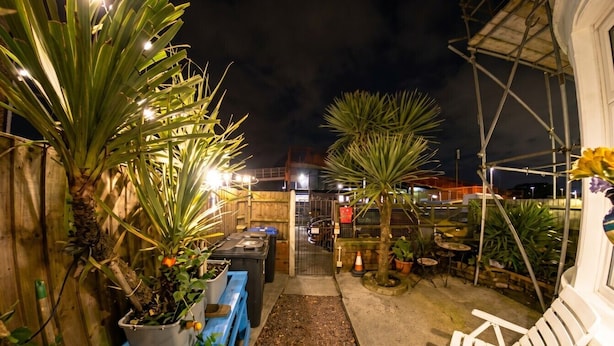 Un espacio al aire libre de noche con plantas en macetas, un banco azul y sillas blancas.