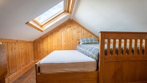 Una habitación de madera con techo inclinado, una cama con colchón y almohadas, y un banco de madera.