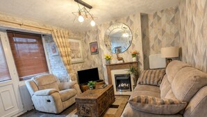 Una sala de estar con sofá, sillón, mesa de centro de madera, chimenea, televisión y objetos decorativos.