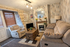 Una sala de estar con sofá, sillón, mesa de centro de madera, chimenea, televisión y objetos decorativos.