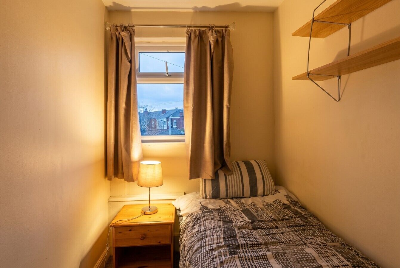 Une petite chambre avec un lit simple, une table de chevet avec une lampe, une fenêtre avec des rideaux et une étagère au-dessus du lit.