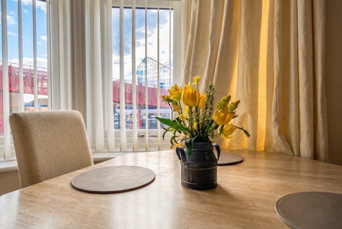 A dining table with a vase of yellow flowers, a chair, and a view of a Ferris wheel through a window with white curtains.