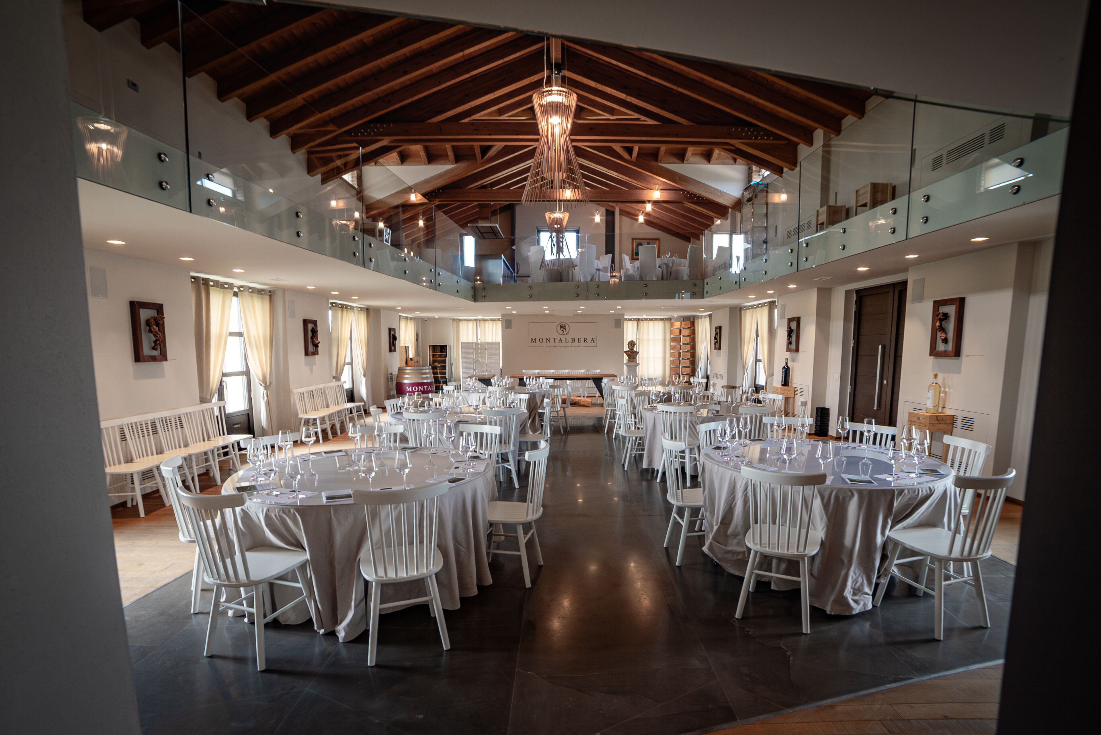 A spacious hall with multiple round tables and chairs, a high ceiling with wooden beams, and a balcony overlooking the dining area.