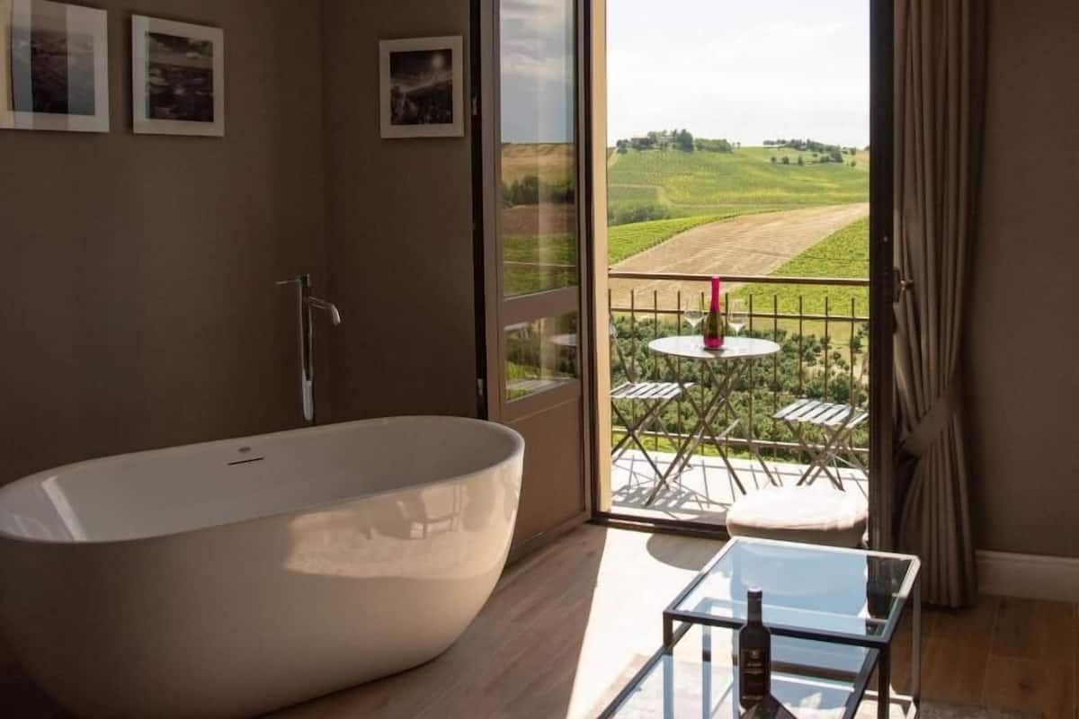 Ein modernes Badezimmer mit einer freistehenden Badewanne, einem Glastisch mit einer Flasche sowie einem Balkon mit Tisch und Stühlen.