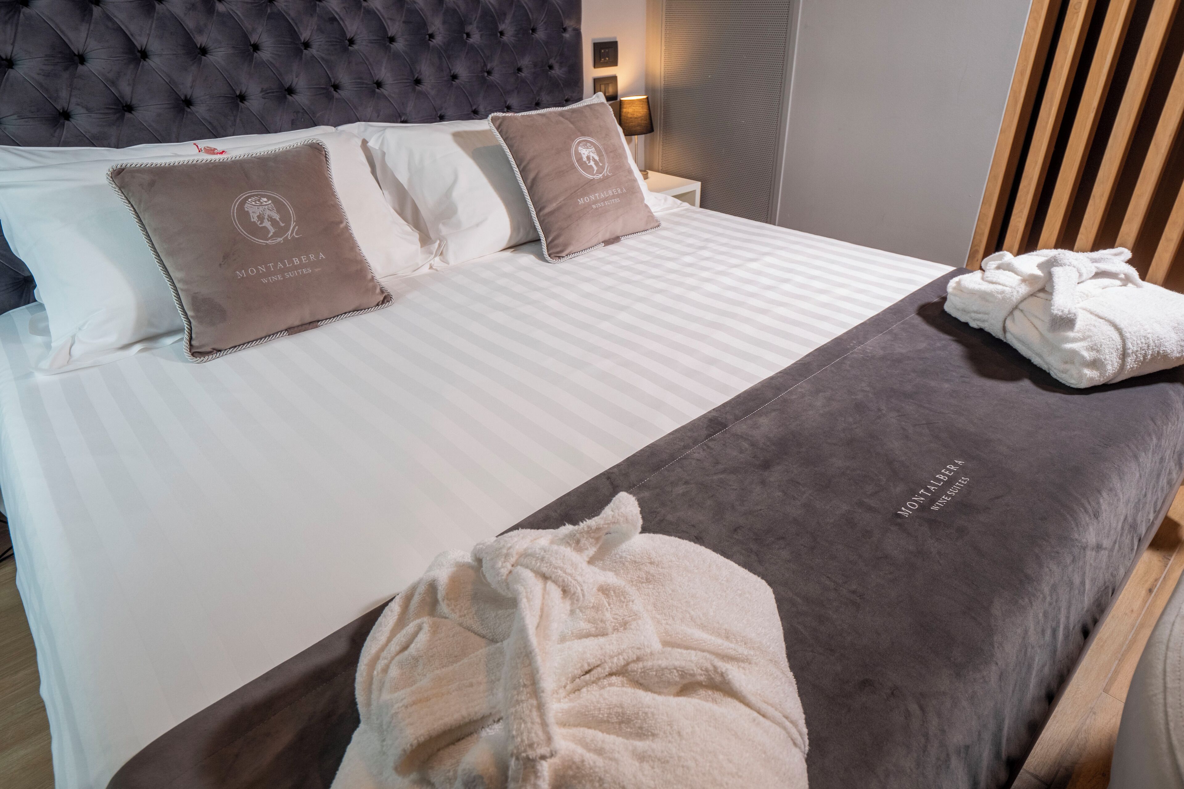 A neatly made bed with a grey headboard, two pillows with a logo, a white blanket, and a rolled white towel on the bed.