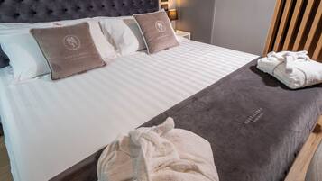 A neatly made bed with a grey headboard, two pillows with a logo, a white blanket, and a rolled white towel on the bed.