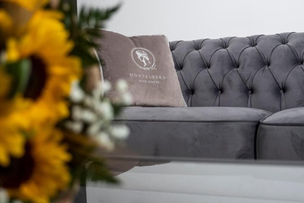A grey tufted sofa with a pillow displaying 'Montalbera Wine Suites' logo.