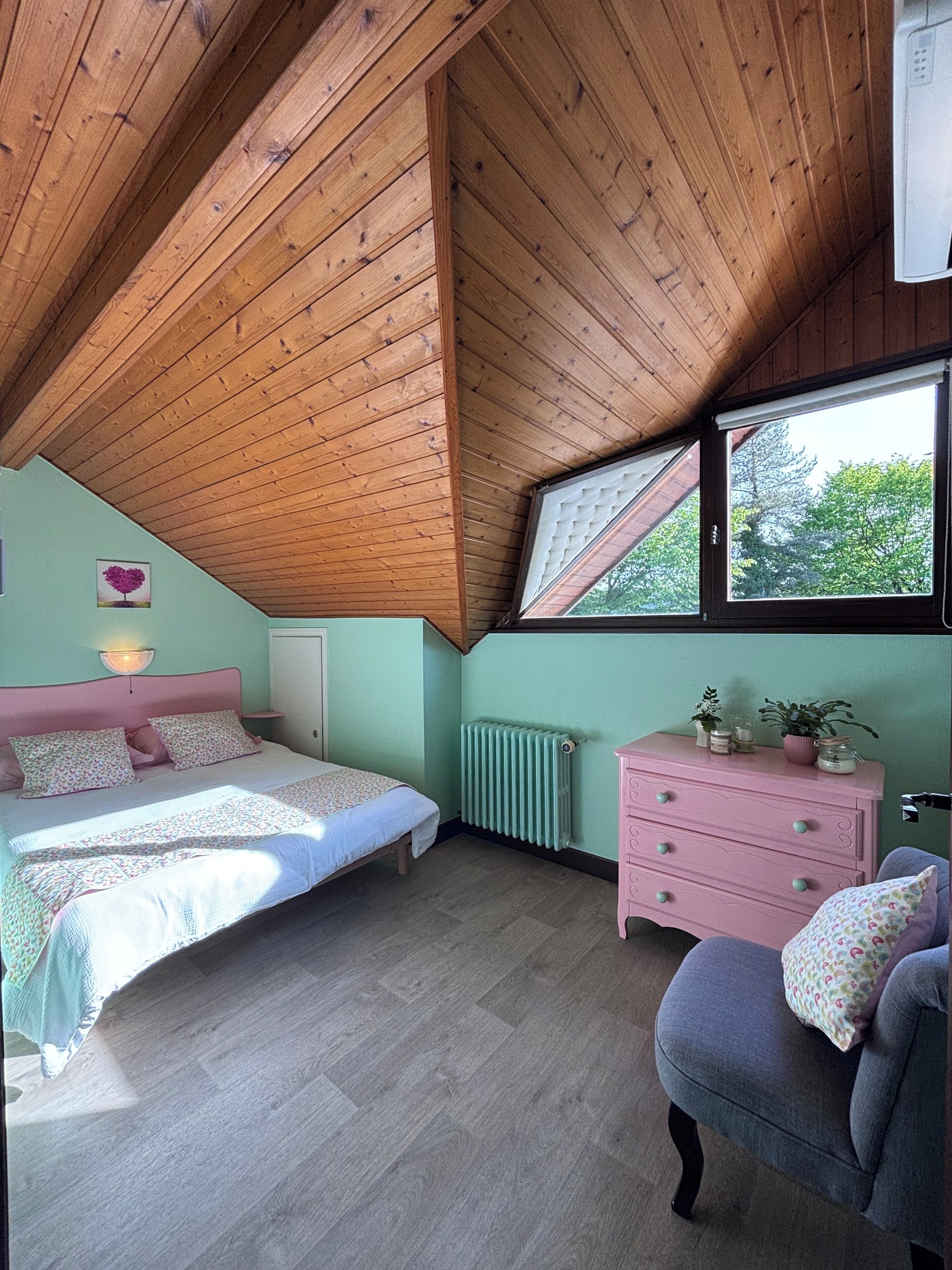 Ein Schlafzimmer mit schräg verlaufender Holzbalkendecke, einem Bett mit rosa Kopfteil, einem Schminktisch und einem Fenster mit Blick auf Bäume.