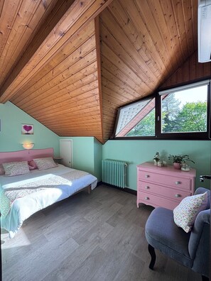Een slaapkamer met een schuin houten plafond, een bed met een roze hoofdeinde, een dressoir en een raam met uitzicht op bomen.