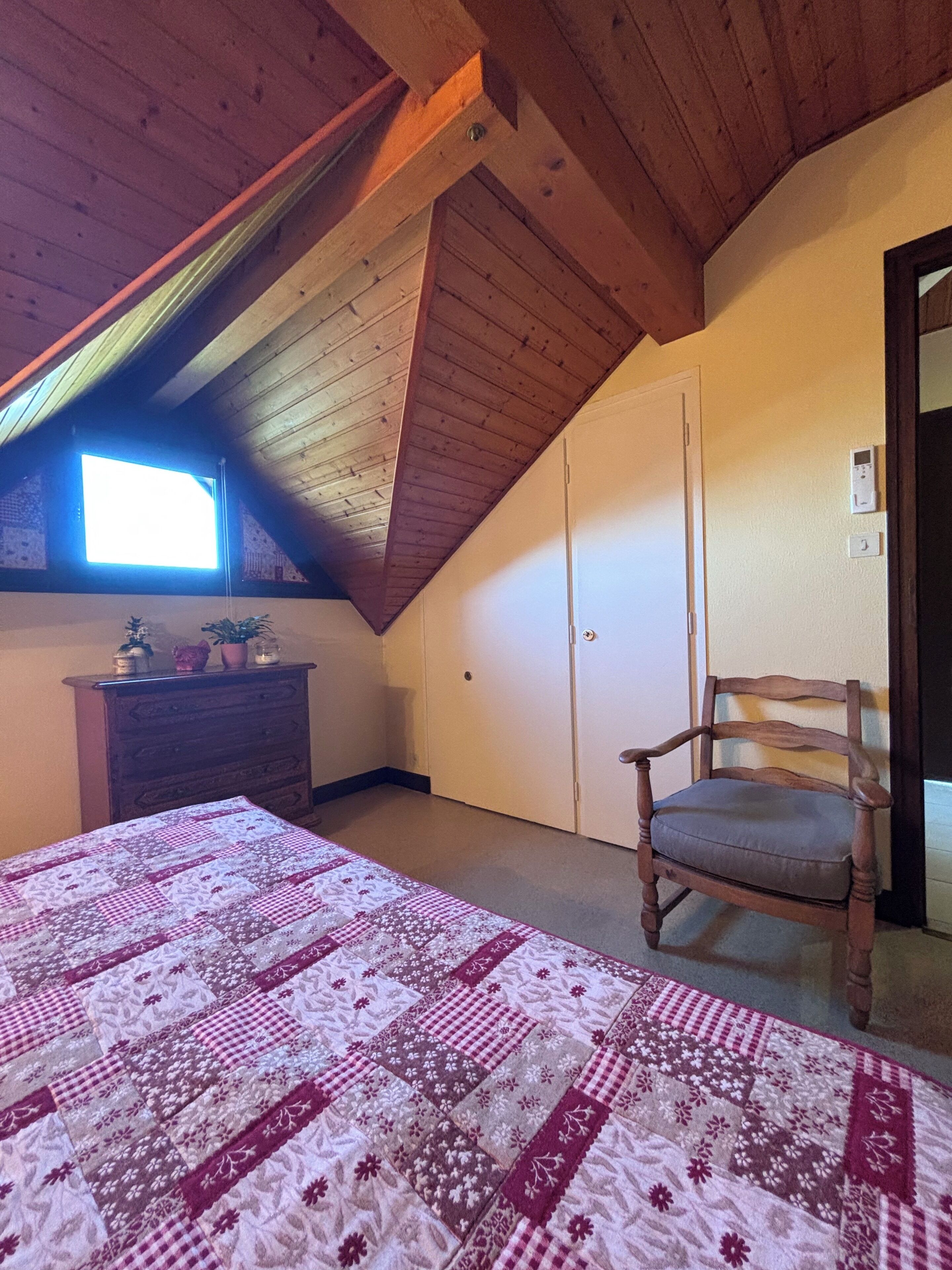 Un dormitorio con techo inclinado, un aparador de madera, una silla, un colcha y una ventana.