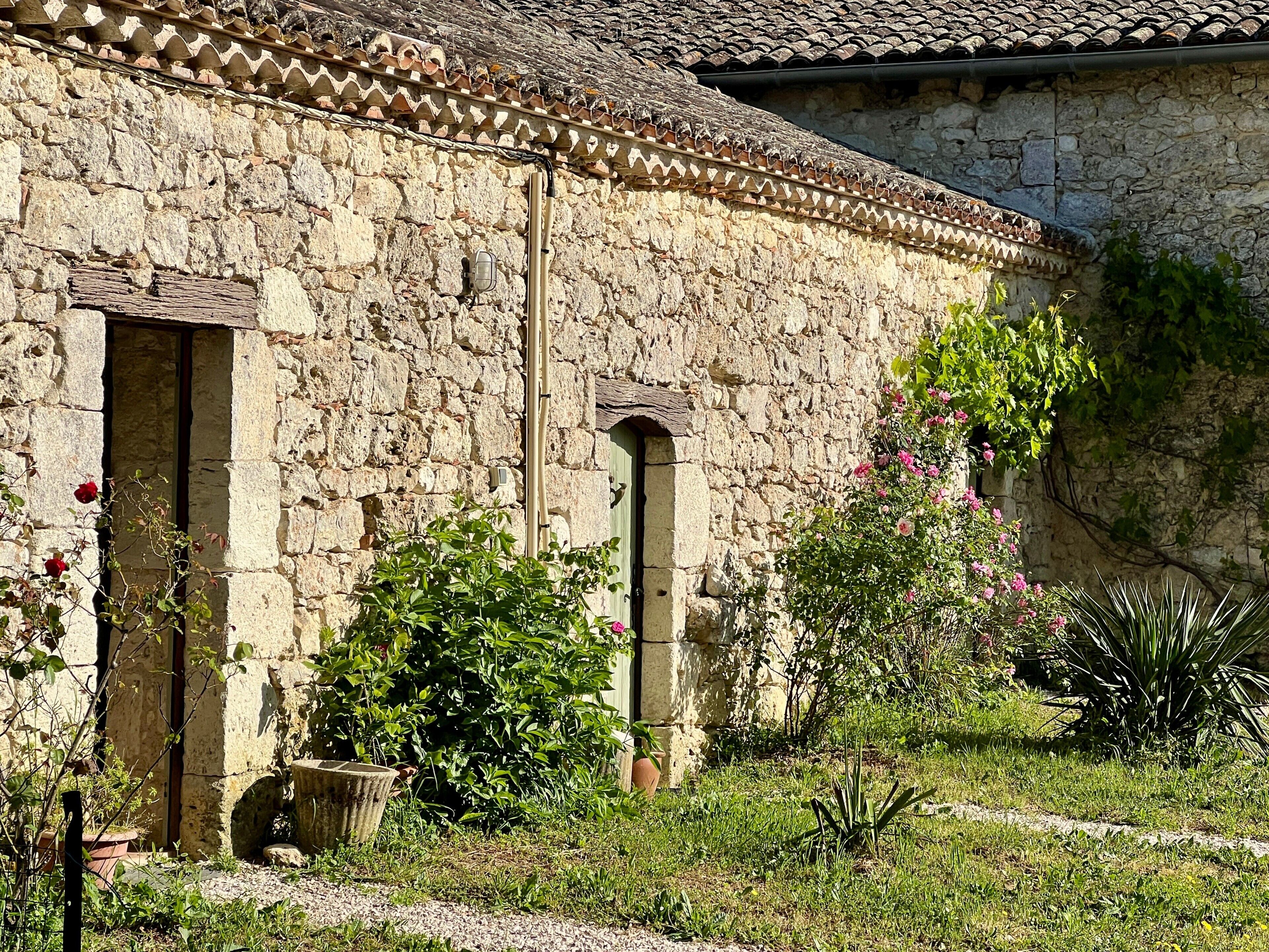 Ein Gebäude aus Stein mit einem Ziegeldach, einer Tür und einem Fenster. Ein Garten mit verschiedenen Pflanzen und Blumen ist vorhanden.