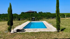 Una piscina rectangular con una estructura oscura en el techo al fondo, rodeada de césped y dos árboles altos.