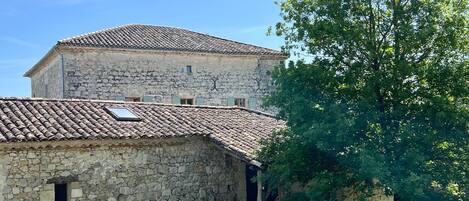 Un edificio de piedra con techo de tejas, una ventana pequeña y un árbol en primer plano.