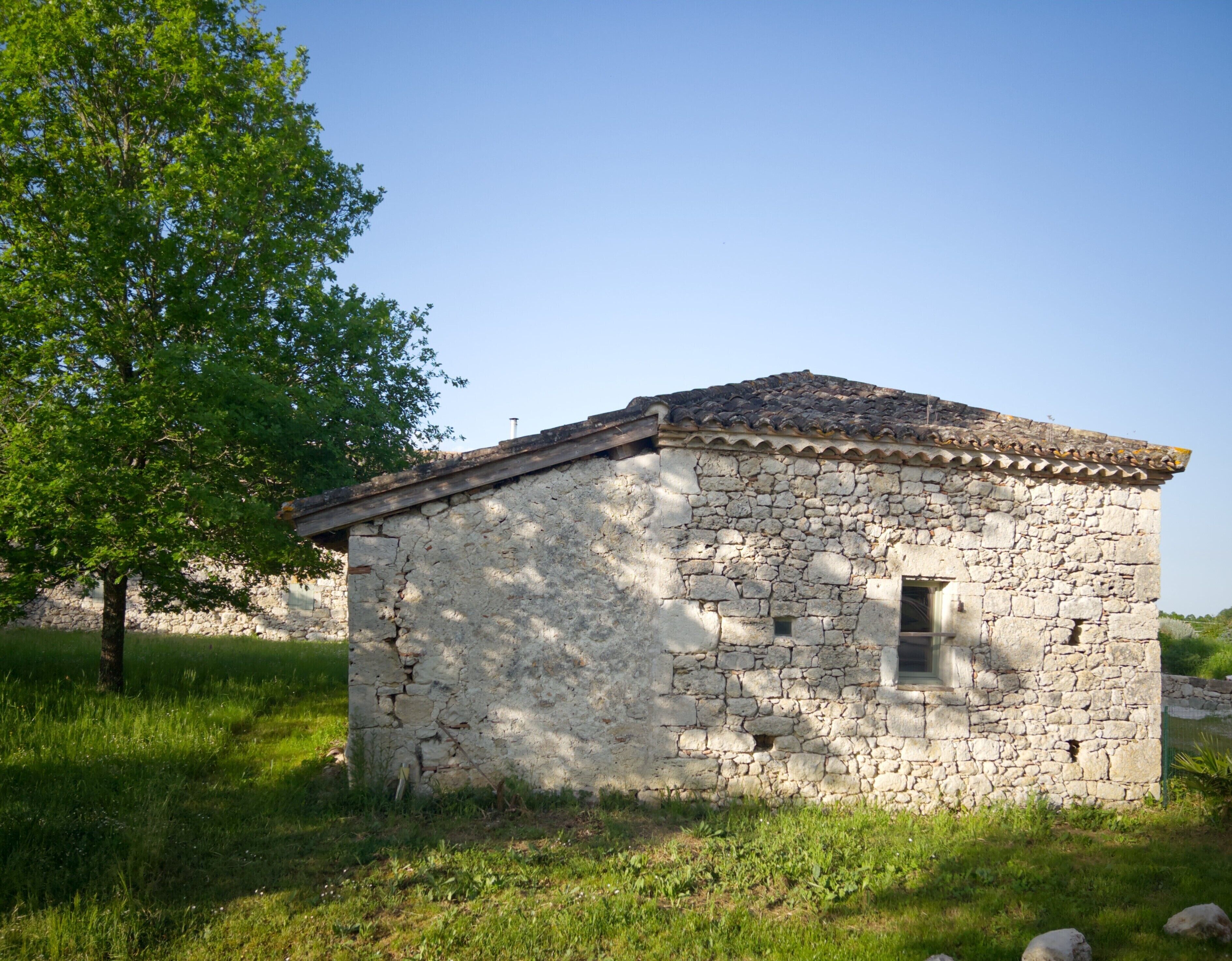 A small stone building with a tiled roof, surrounded by grass and a tree.