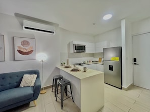 A modern kitchen with white cabinets, a stainless steel refrigerator, a microwave, and a small island. There is a blue sofa with a cushion and two stools in the living area. - Modern Apartment at PH Living 73 San Francisco (Panamá)