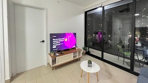 Un soggiorno moderno con televisore a schermo piatto, un tavolino bianco e un'ampia finestra vista città.