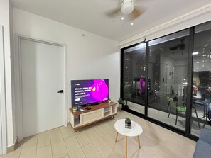 Ein modernes Wohnzimmer mit Flachbildfernseher, einem kleinen weißen Tisch und einem großen Fenster mit Blick auf die Stadt.
