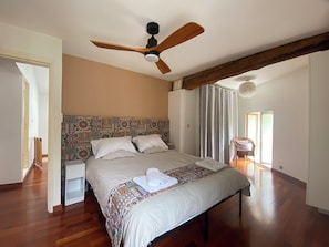 Un dormitori amb llit, ventilador de sostre, capçalera amb dibuixos, una cadira i una porta que dona a un altre espai.