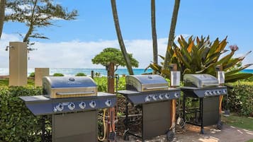 Three barbecue grills with controls, positioned outdoors with a view of the ocean and palm trees.