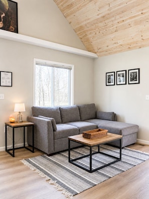 Ein modernes Wohnzimmer mit einer grauen Couch, einem Holztisch, einem kleinen Beistelltisch mit einer Lampe und einem großen Fenster.