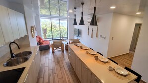A modern kitchen with a central island, a dining area with chairs, and a large window providing a view of greenery. - Hotel Macondo Quinta Avenida by Ruha (Playa del Carmen)