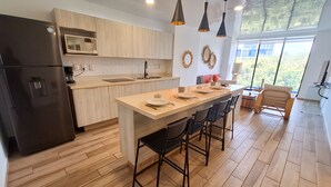 A modern kitchen with a central island, bar stools, a refrigerator, and a large window providing a view of the outdoors. - Hotel Macondo Quinta Avenida by Ruha (Playa del Carmen)