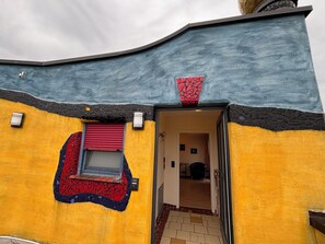 A building with a unique mosaic design, a red shuttered window, and a door leading to an interior space. - Hirn Winery & HUNDERTWASSER articles in the wine paradise (Eisenheim)
