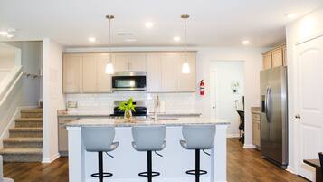 A modern kitchen with a central island, bar stools, stainless steel appliances, and a staircase.