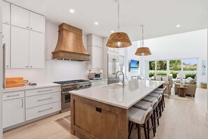 A modern kitchen with white cabinetry, a large island with bar stools, a stainless steel stove, and a wooden range hood. - New Luxury Retreat - Resort Pool - Walk to Beach (Naples)
