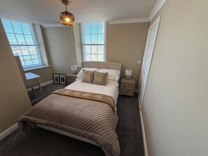 Une chambre à coucher comprenant un lit, des tables de chevet, un bureau et deux fenêtres équipées de stores.