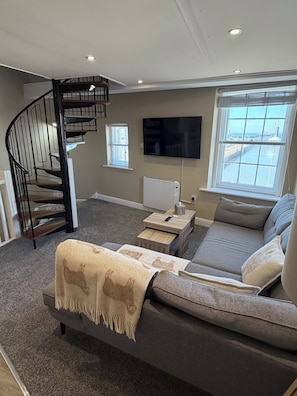 Un salon avec un canapé gris, une table basse, un téléviseur, une cheminée et un escalier en colimaçon.