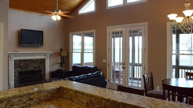 A living room with a stone fireplace, a ceiling fan, a television mounted above it, a dark-colored sofa, and a dining area with a table and chairs.