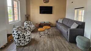 Un salon avec un canapé gris, des fauteuils à motifs, une table basse en bois, un téléviseur fixé au mur et une fenêtre donnant sur l’extérieur.