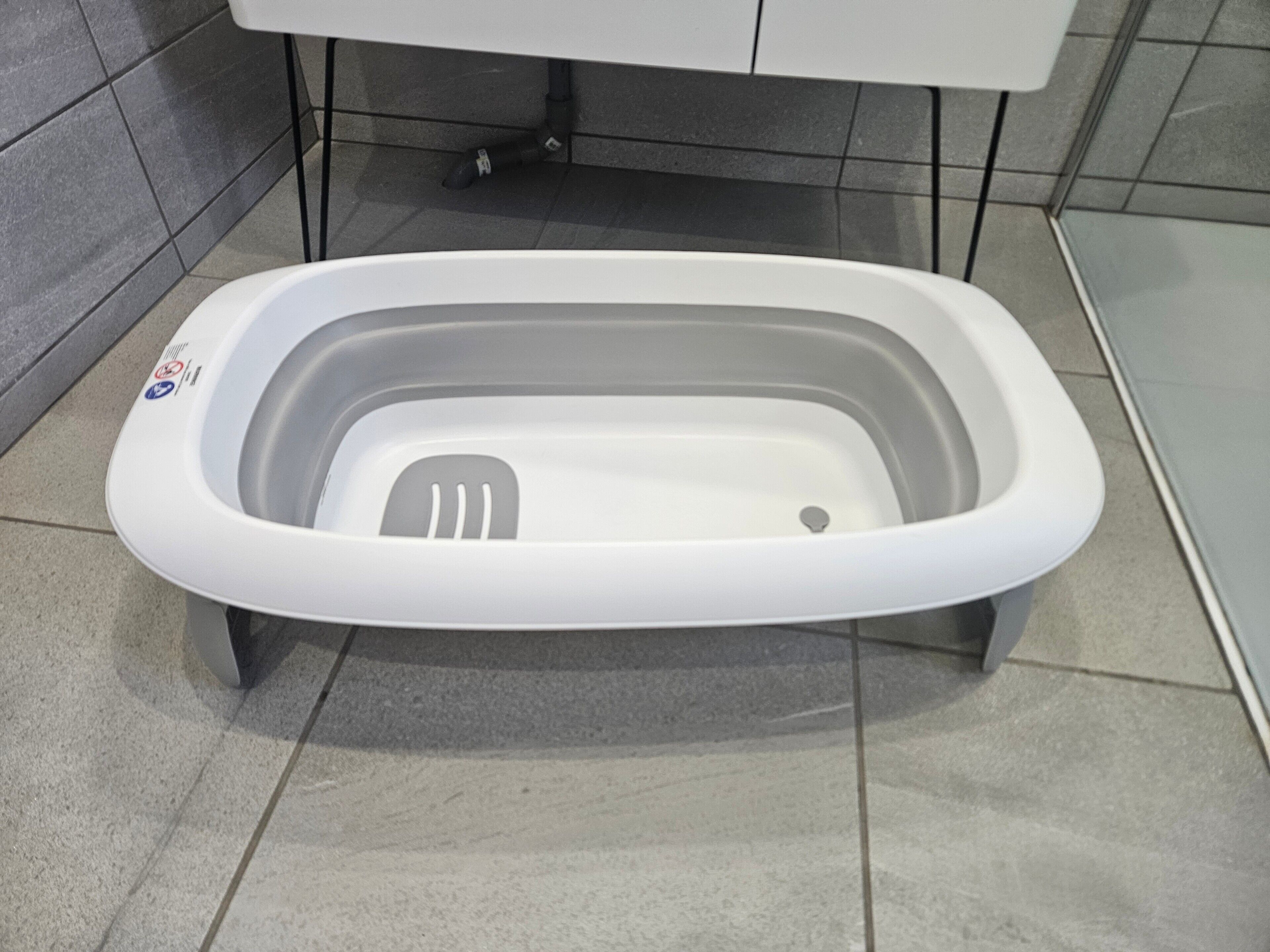 Een witte, verplaatsbare toiletcabine met voetpedaal, op een betegelde vloer in een badkamer met grijze tegels en een witte wasbak op de achtergrond.