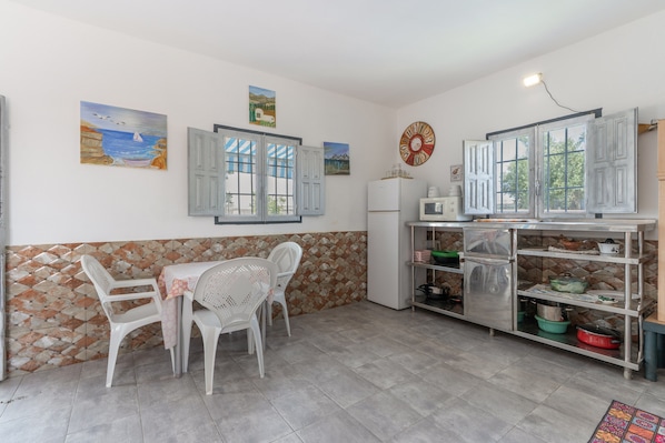 A kitchen with a dining table, chairs, a refrigerator, a microwave, and a shelf with kitchenware.