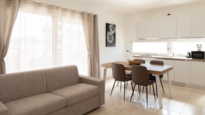 A living room with a sofa, dining table, chairs, and a kitchen area with cabinets and a microwave. - La Dimora Molara by Wonderful Italy (Olbia)