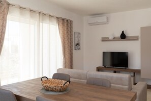 A modern living room with a wooden dining table, beige sofas, a television on a stand, and a window with sheer curtains. - La Dimora Tavolara by Wonderful Italy (Olbia)