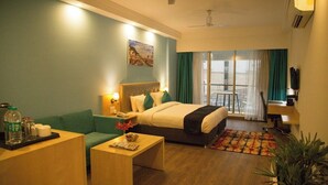 A hotel room with a bed, a sofa, a desk, a chair, a plant, and a view of the outside. - Saramax Inn (Roorkee)