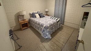 Un dormitorio con cama, dos mesitas de noche, un estante en la pared, una puerta con manija y una lámpara.