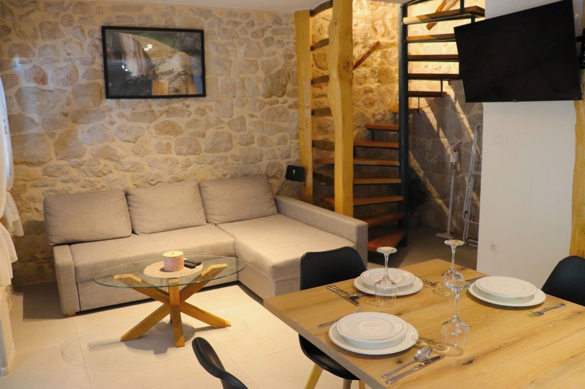 Un espacio de estar con un sofá, una mesa de centro de vidrio, una mesa de comedor con platos y copas de vino, una pared de piedra y una escalera de madera.