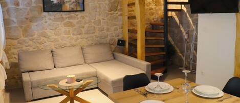 Un espacio de estar con un sofá, una mesa de centro de vidrio, una mesa de comedor con platos y copas de vino, una pared de piedra y una escalera de madera.