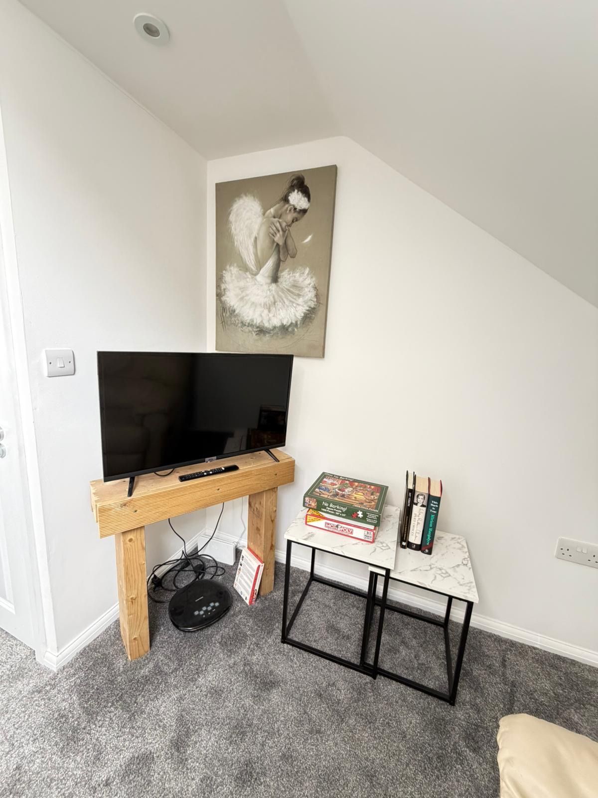 Ein Zimmer mit Fernseher auf einem Holzboden, einem kleinen Tisch mit Büchern und einem Gesellschaftsspiel sowie einem Bild an der Wand.