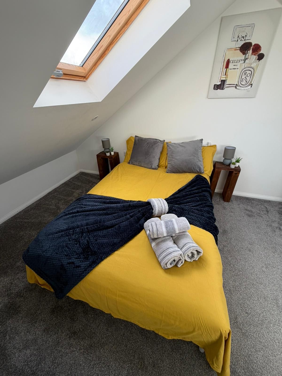 Ein Schlafzimmer mit schrägem Dach, einem Bett mit gelber Bettwäsche, einer dunkelblauen Decke und gerollten Handtüchern. Auf einem Nachttisch mit Lampe und kleiner Pflanze.