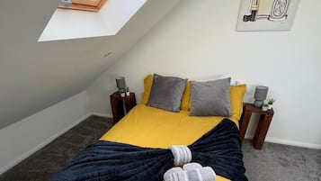 Ein Schlafzimmer mit schrägem Dach, einem Bett mit gelber Bettwäsche, einer dunkelblauen Decke und gerollten Handtüchern. Auf einem Nachttisch mit Lampe und kleiner Pflanze.