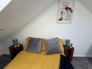 Un dormitorio con techo inclinado, cama con ropa de cama amarilla, dos almohadas grises y un cuadro en la pared.