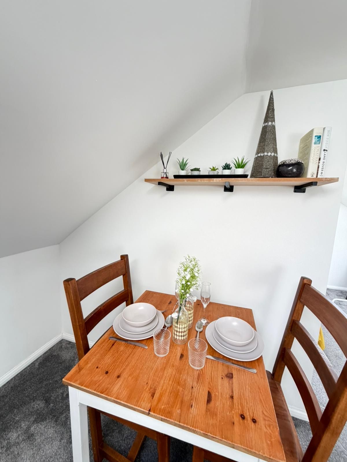 A dining area with a wooden table set for four, chairs, a small shelf with decorative items, and a book on the wall.