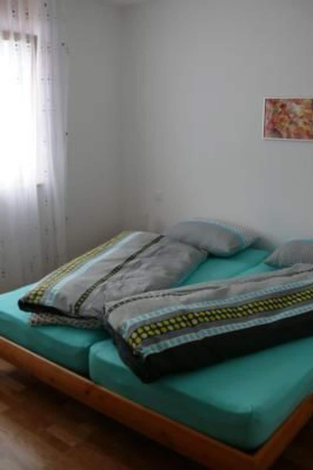 Un lit simple avec un matelas de couleur turquoise et deux oreillers, une tête de lit en bois, une fenêtre avec des rideaux et un petit tableau décoratif au mur.