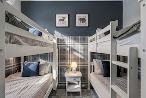 Quarto com beliches, dois quadros de animais na parede, uma mesinha com abajur e uma janela com cortina xadrez.