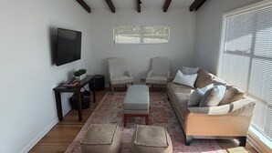 Un salon comprenant un canapé, deux fauteuils, une table basse, un téléviseur et une fenêtre avec des stores.
