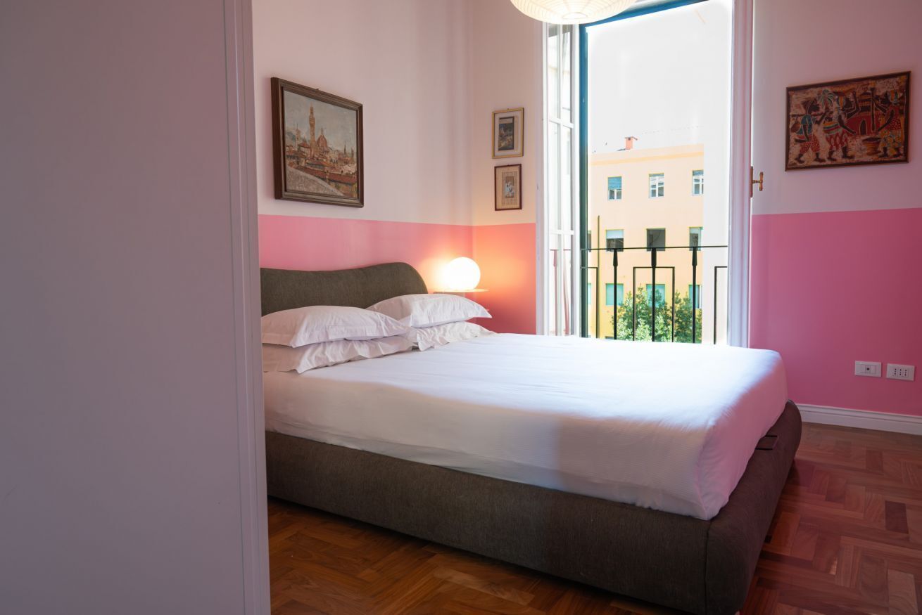 A bedroom with a bed, a nightstand, a window with a view of buildings, and a door leading to another room.